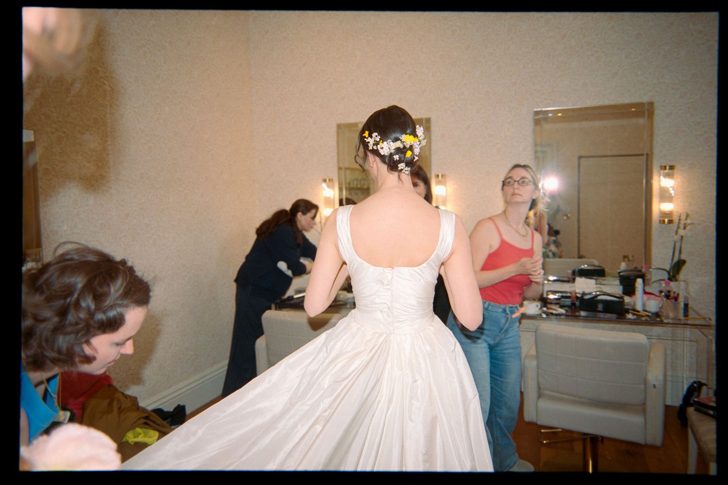 A bride in a white gown with a floral hair accessory stands in a room while two women prepare nearby on her wedding day.