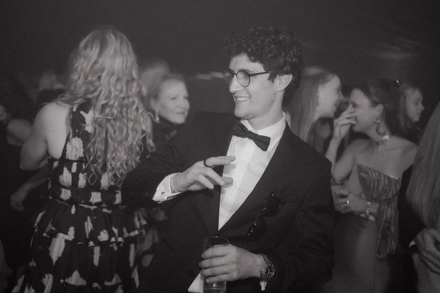 Black and white photo of a smiling curly-haired man in glasses, tuxedo and bow tie gesturing while guests mingle at a formal evening party.