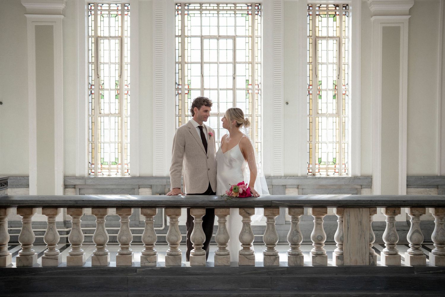 Elegant bride and groom standing together at ornate white balustrade inside a grand venue with tall decorative windows.