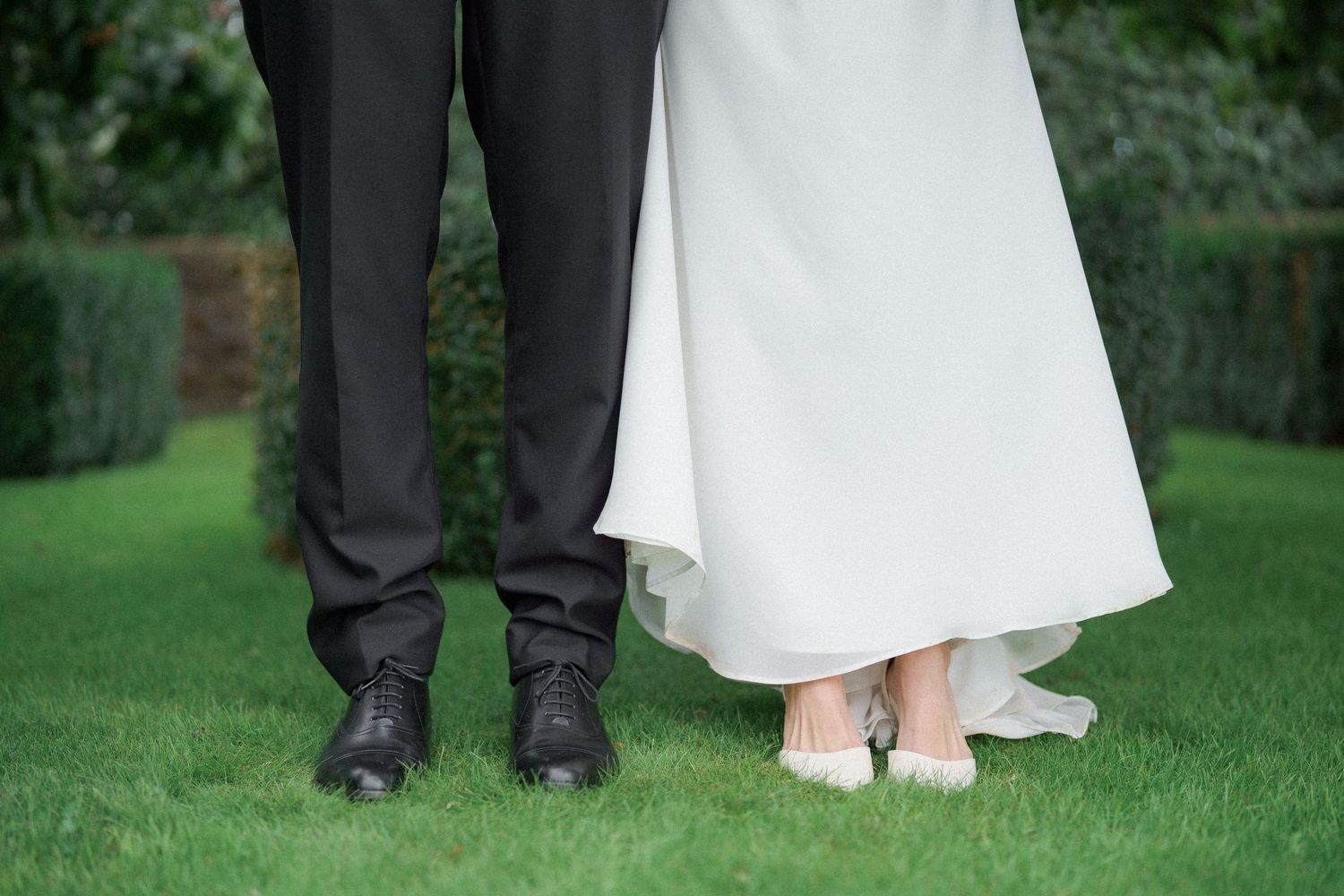 Close-up view of formal dress shoes and white gown hem on green grass at outdoor wedding.