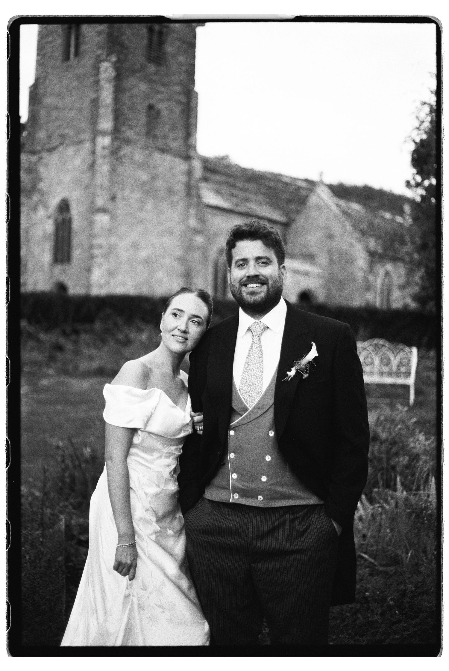Bride and bearded groom pose together outside rustic stone church after wedding.