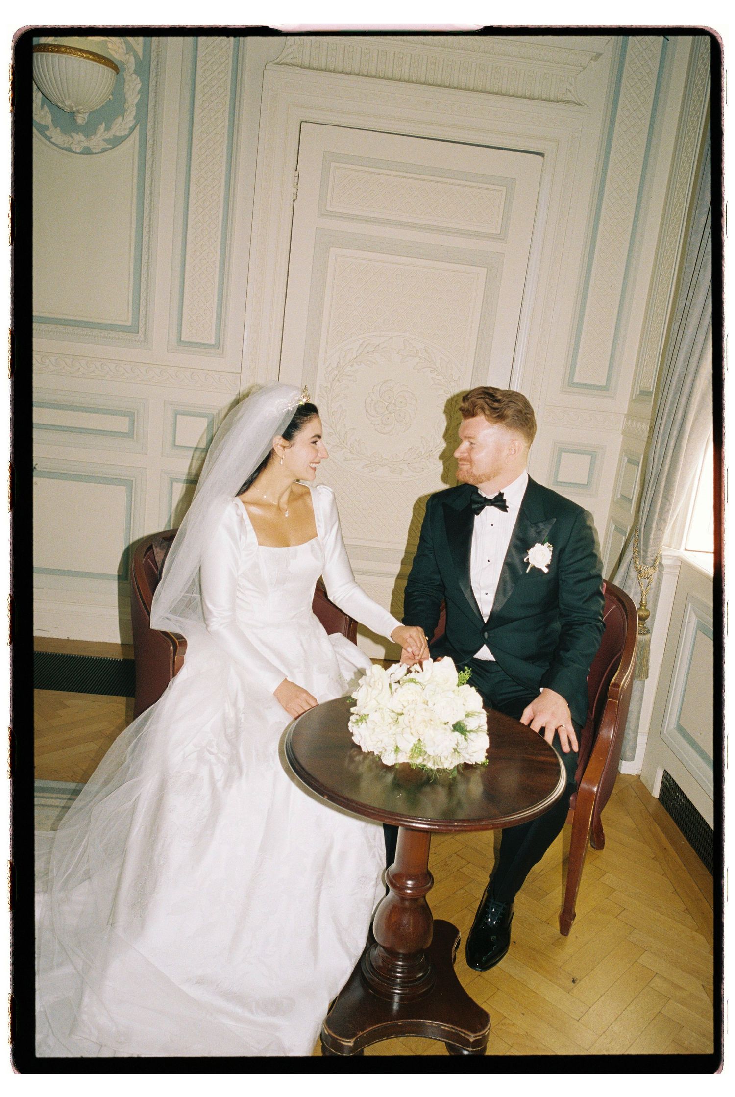 Bride and groom sitting at ornate table in elegant French-style room, sharing a romantic moment.