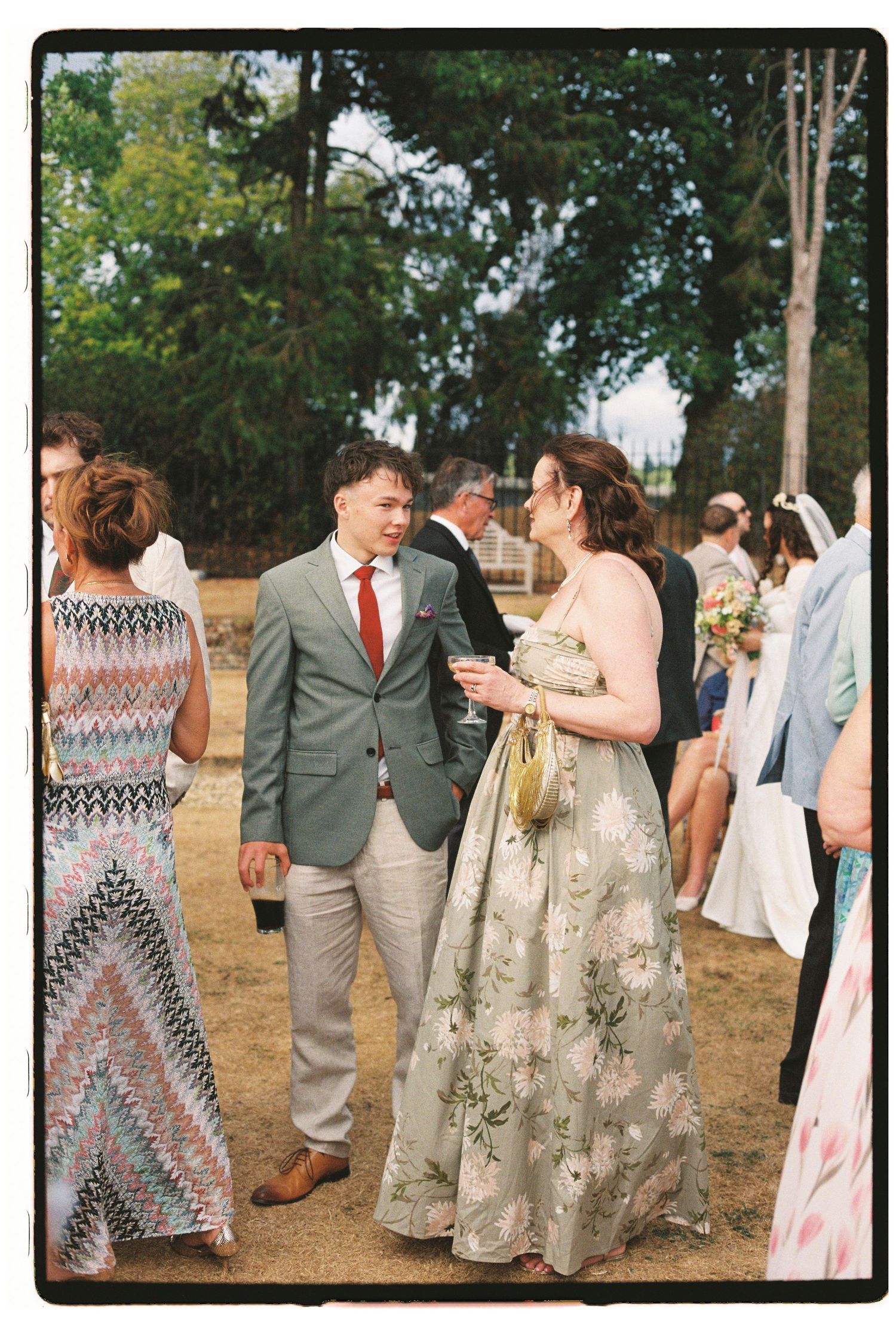 Couple in floral dress and grey suit chat at outdoor wedding reception surrounded by guests.