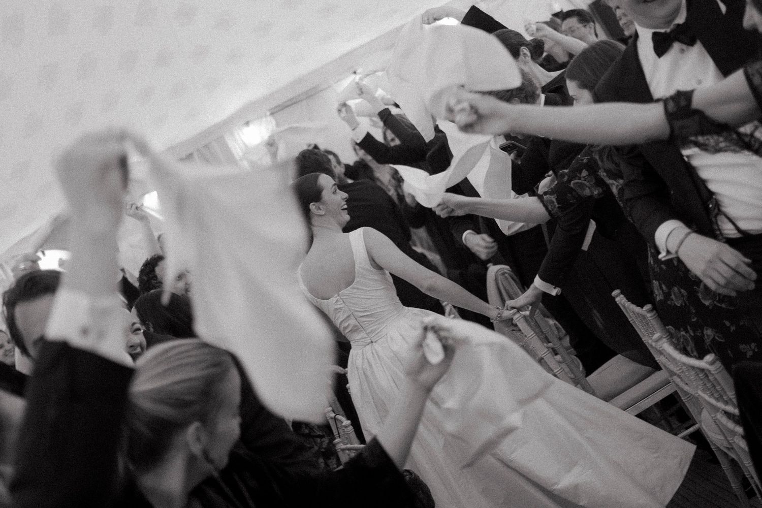 Bride in a white gown dances with guests waving napkins in the air at a lively wedding reception in black and white.