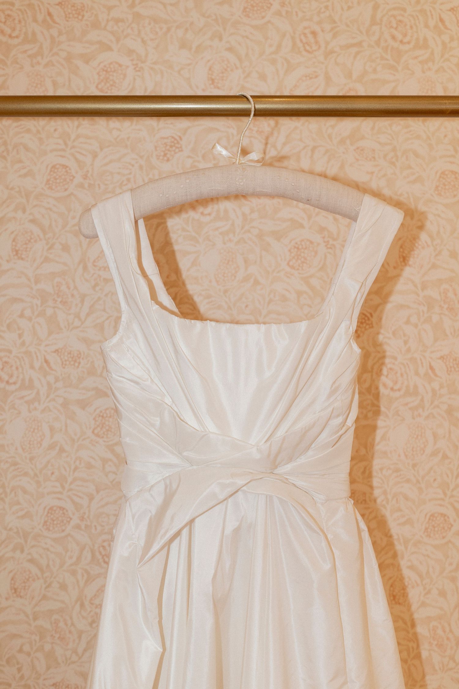 A white sleeveless wedding dress with a gathered bow waist hangs on a white hanger against floral peach wallpaper.