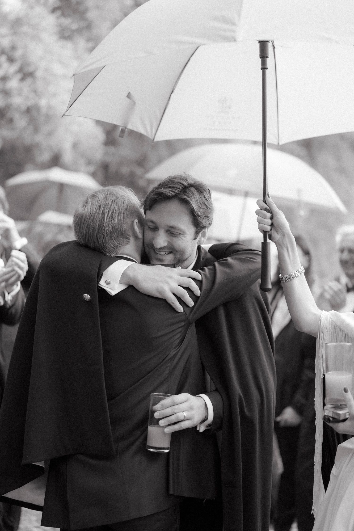 Two men share a warm hug under white umbrellas in black and white at a rainy outdoor wedding celebration.