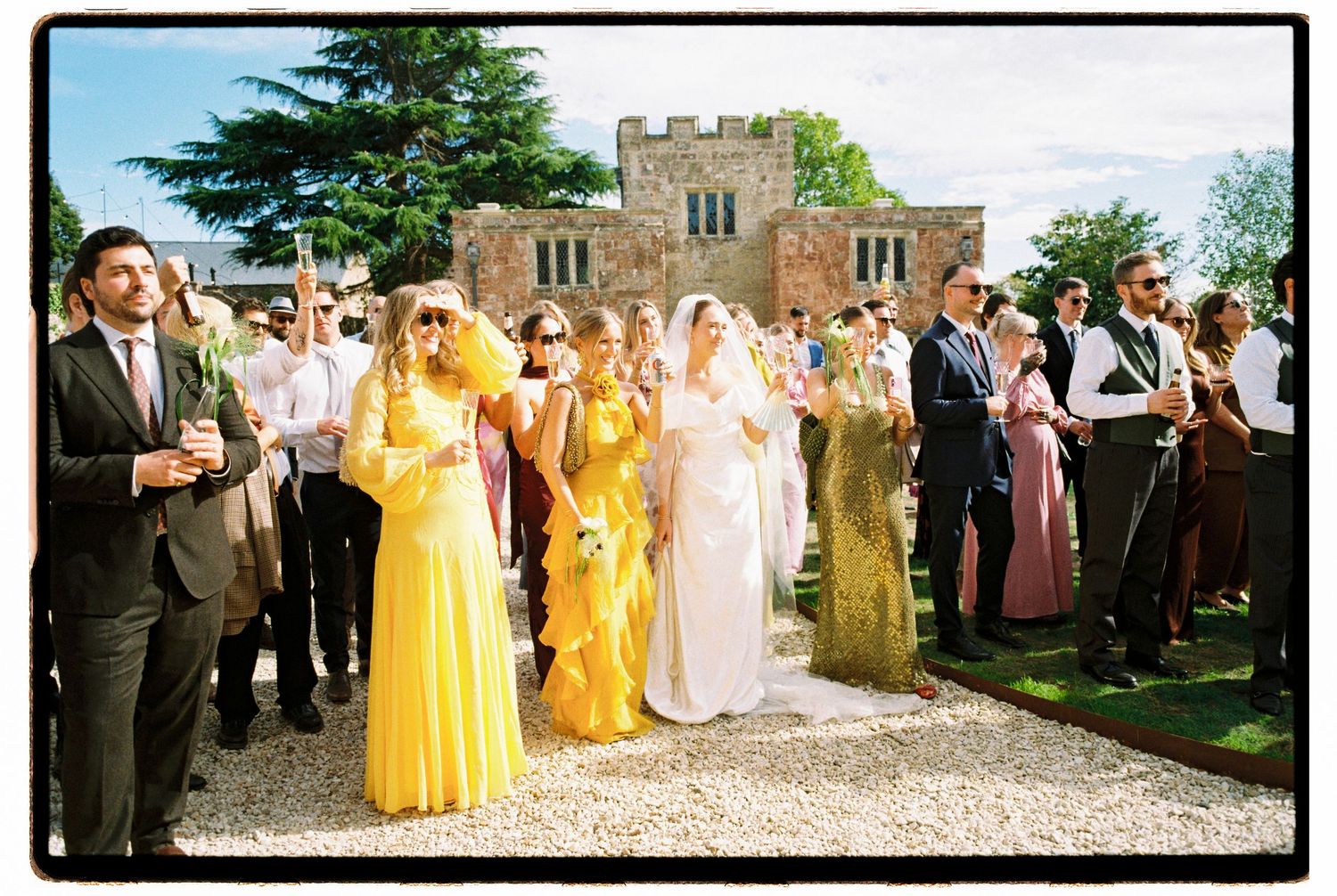 Wedding guests in yellow bridesmaid dresses and formal attire gather outdoors near a historic stone building on a sunny day.