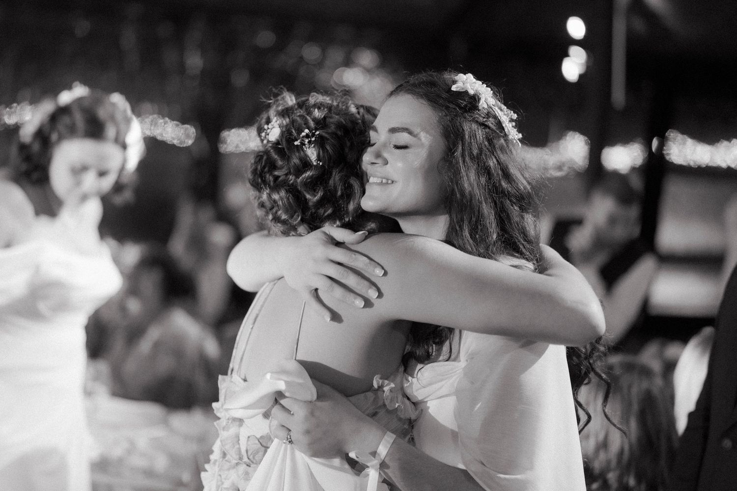 Two women in white dresses share a warm embrace on the wedding dance floor in black and white.