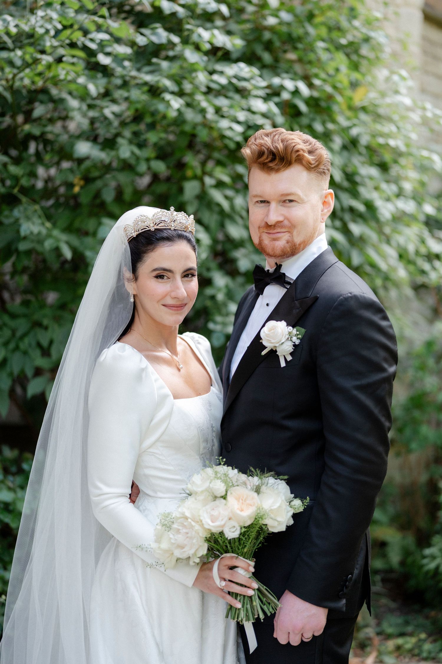 Bride and groom pose outdoors, she holds white bouquet wearing tiara veil, he wears black tuxedo with boutonniere.