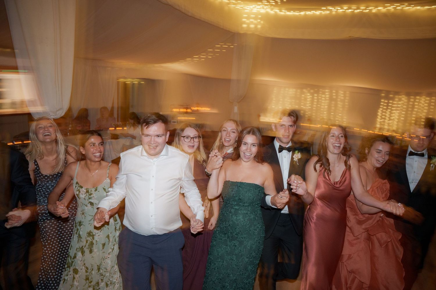 A group of wedding guests dancing together on the dance floor at a reception under warm lighting.
