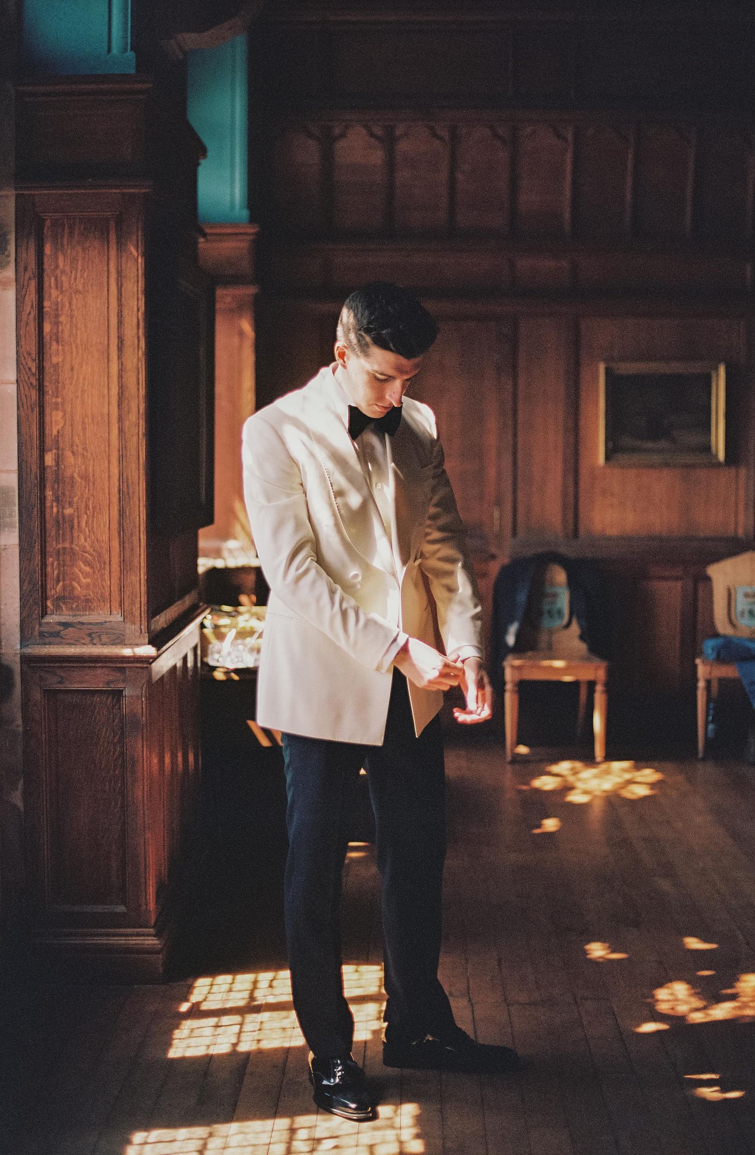 Groom in cream tuxedo jacket and bow tie standing in wood-paneled room with dramatic lighting.