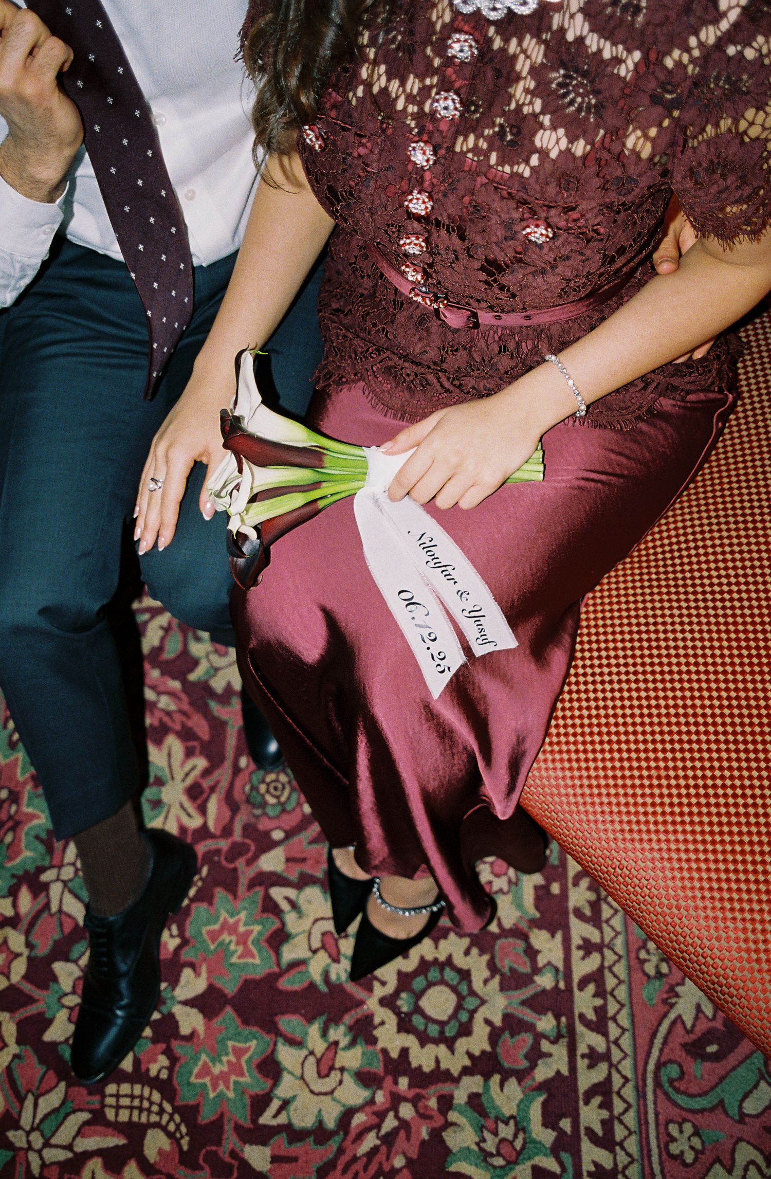 Bride in burgundy lace dress holds white calla lily bouquet with ribbon beside groom in dark suit on ornate carpet.