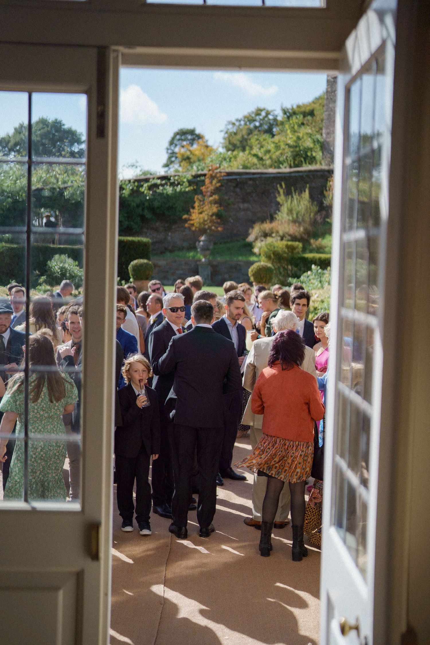 Wedding reception guests mingle in an elegant sunlit room with large windows overlooking a garden landscape.