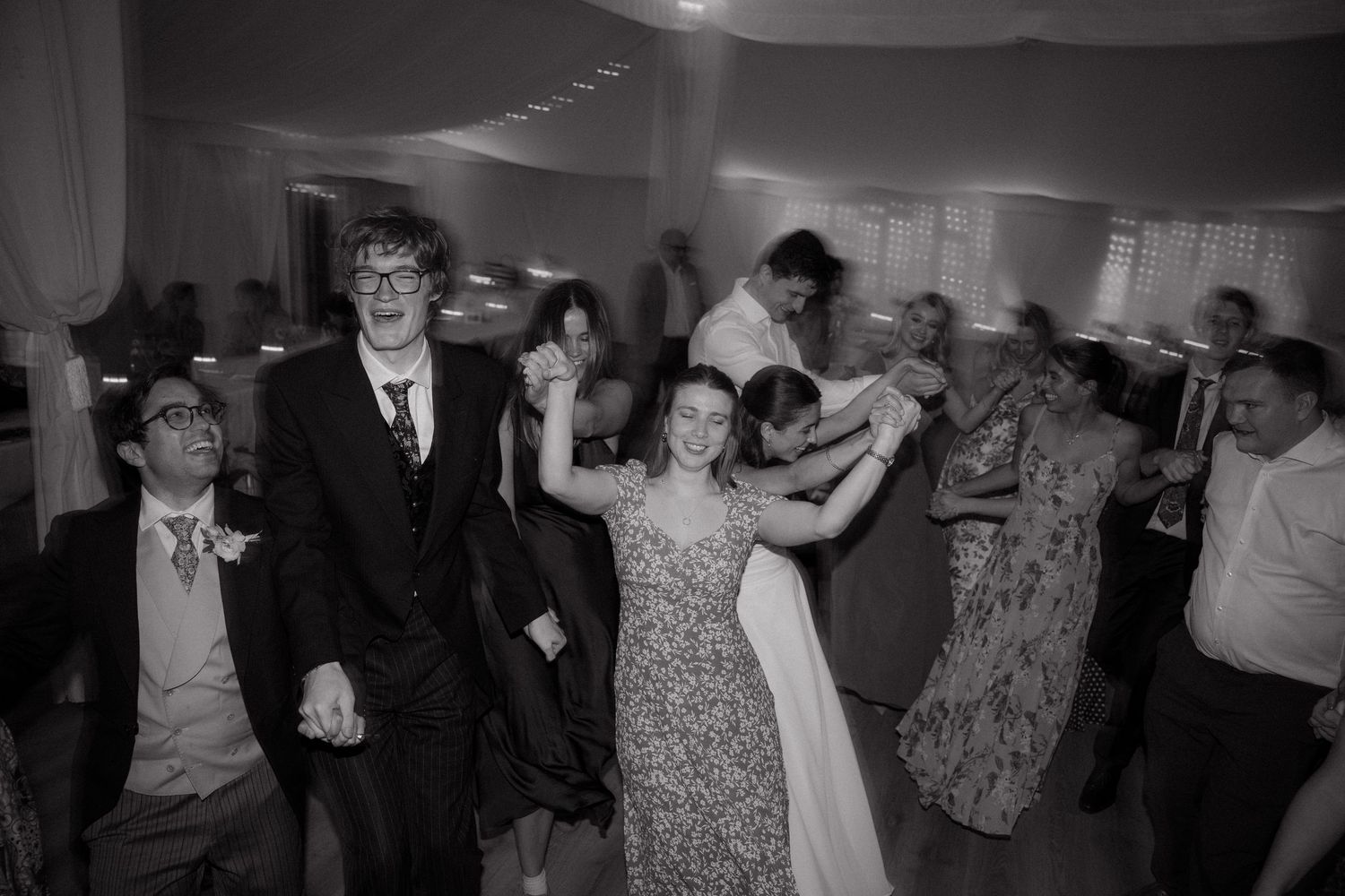 Wedding guests dance and celebrate on a black and white dance floor at an indoor reception venue.