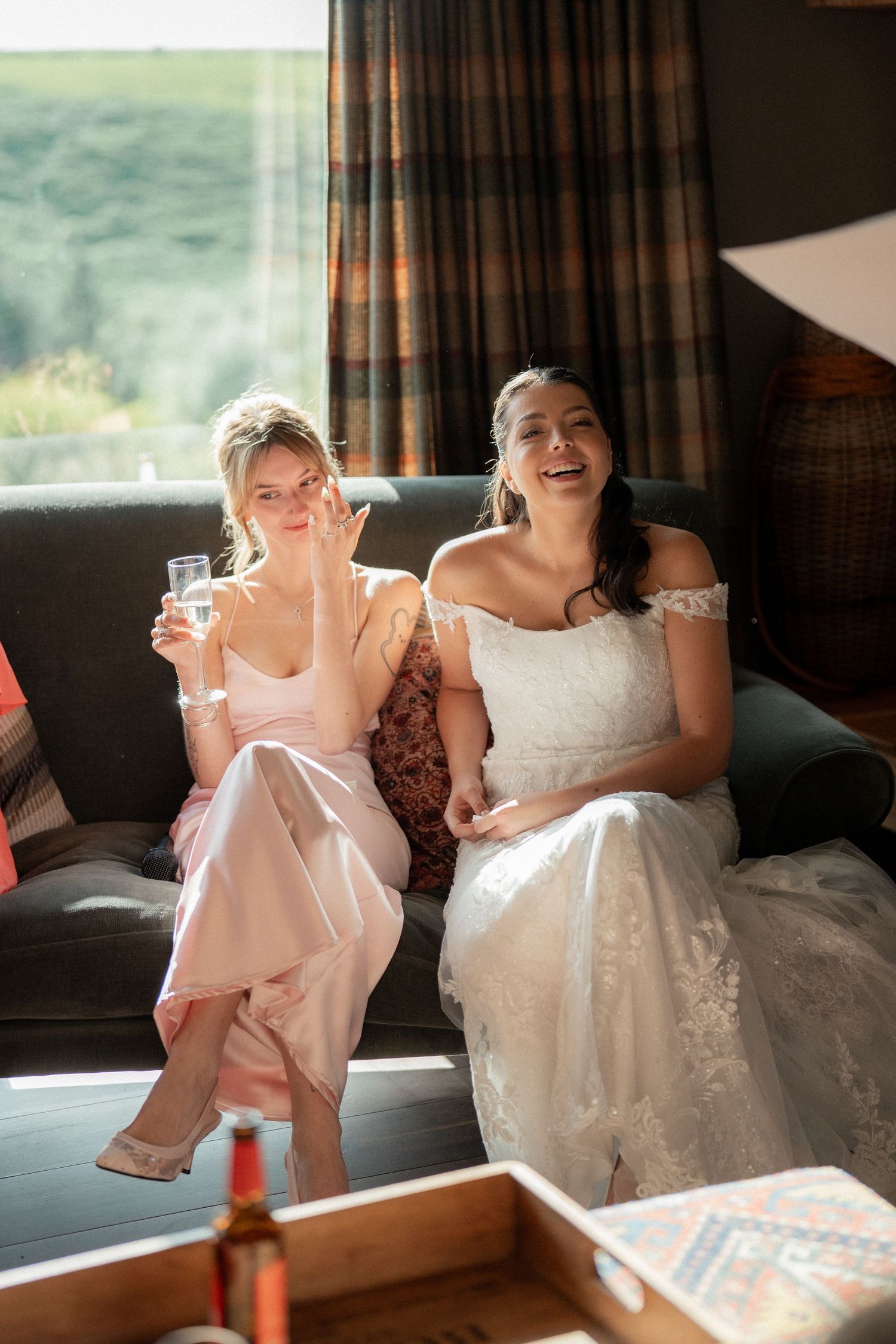 Two women in formal attire share a joyful moment on a couch during a wedding celebration.