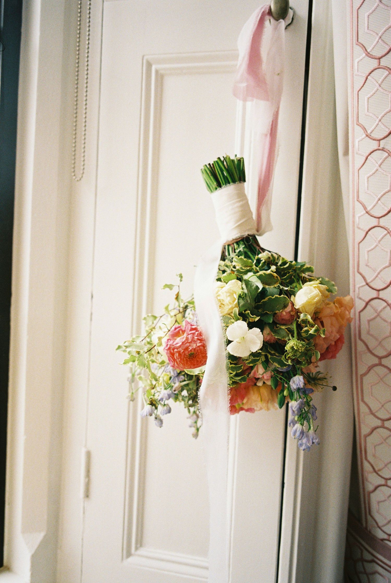 Bridal bouquet of peach roses, white blooms and green foliage tied with ribbon hanging upside down from a white paneled door.
