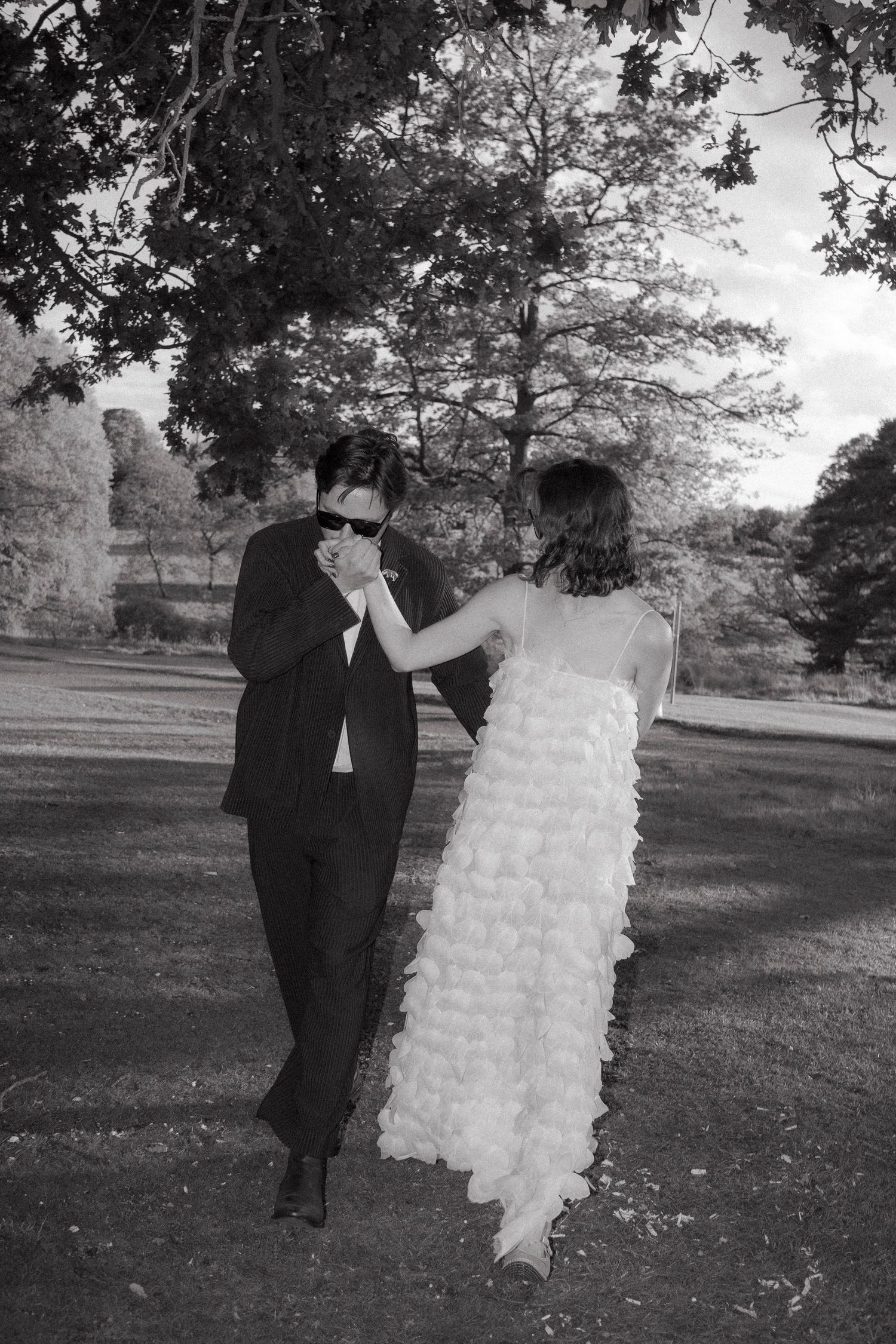 Groom in sunglasses kisses the hand of a bride in a ruffled white gown as they walk across a lawn beneath tall trees.