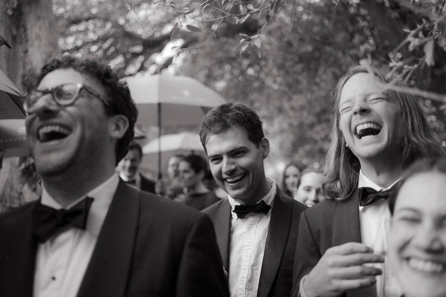Black and white photo of laughing groomsmen and guests in bow ties enjoying a joyful moment outdoors at a wedding.