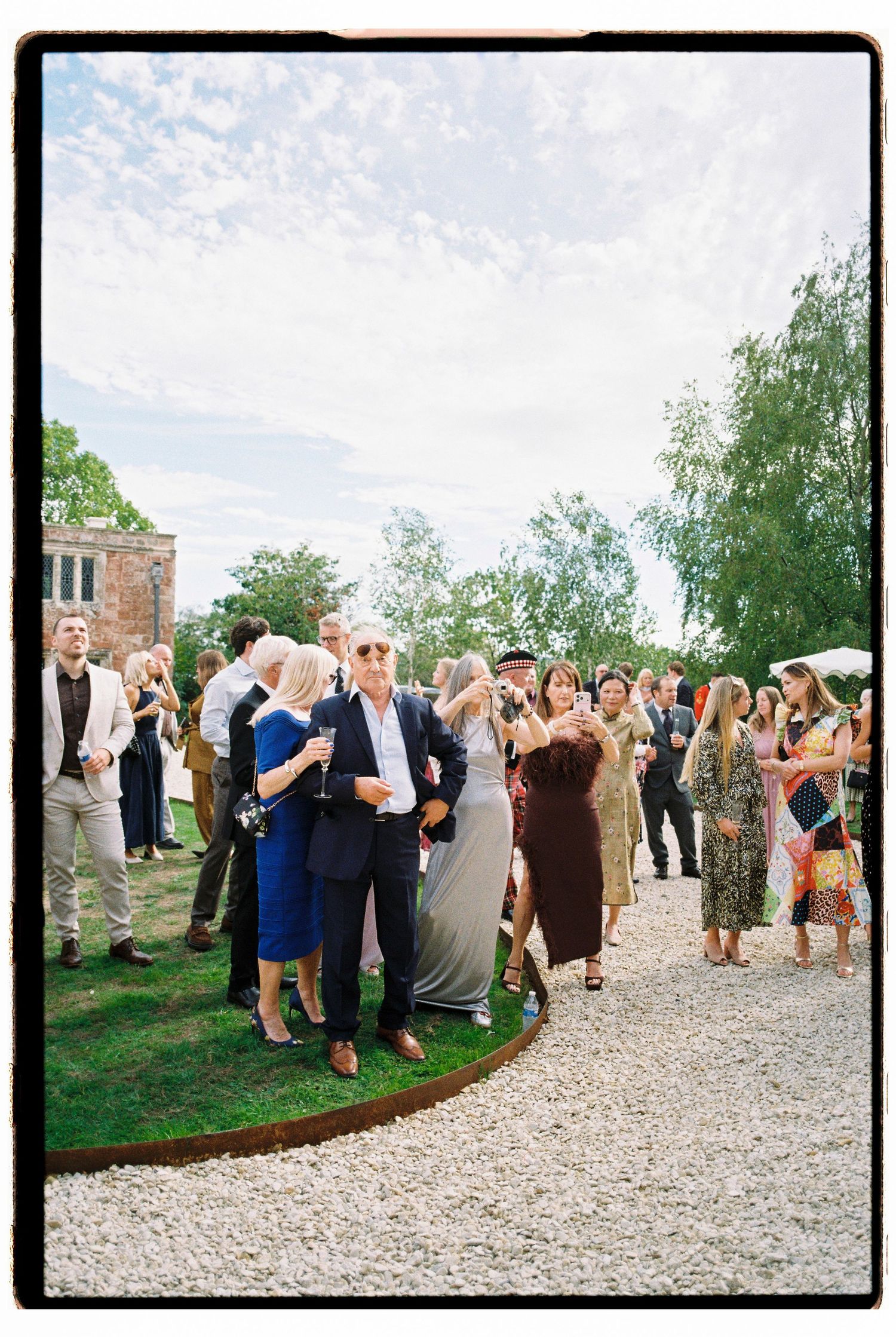 Wedding guests mingle outdoors on sunny day at elegant countryside estate reception venue.