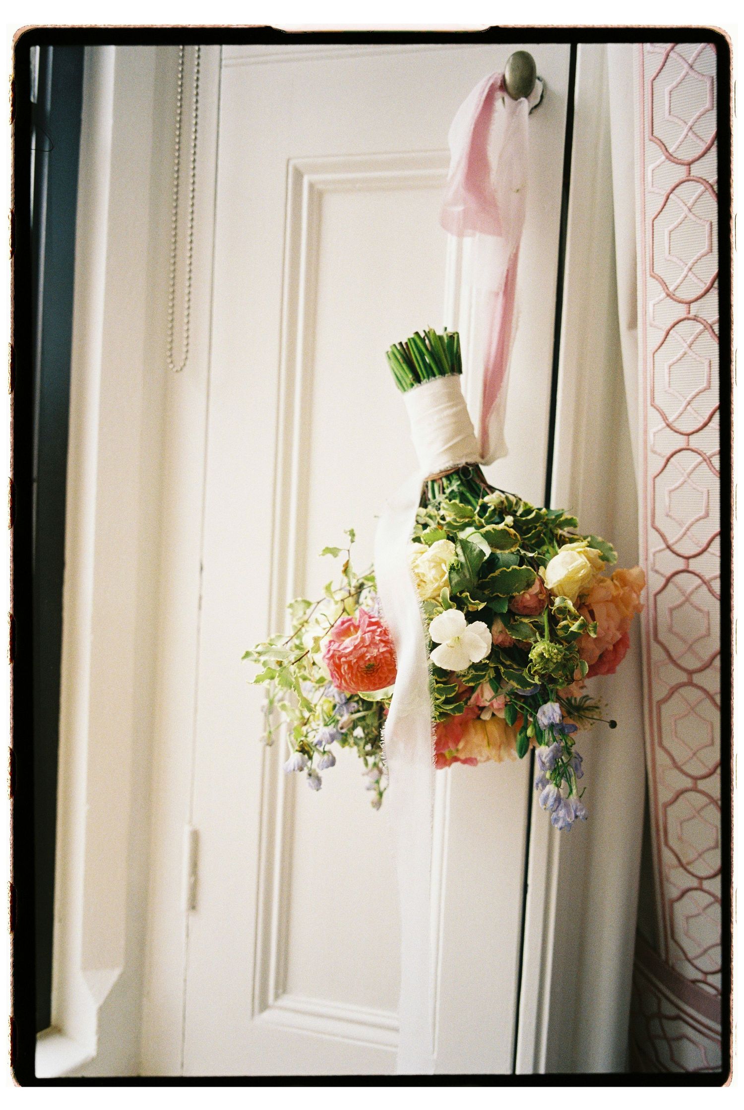 A floral bouquet with pink and white flowers hangs from a white door with a pink ribbon.