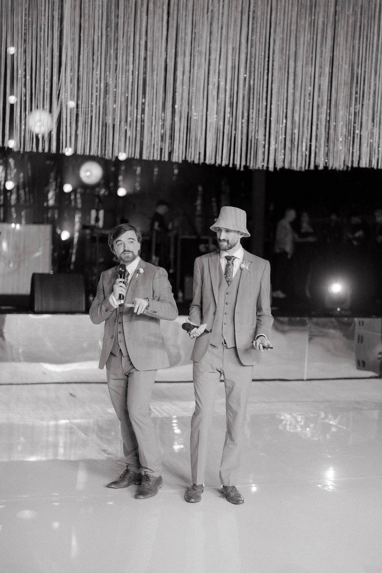 Two grooms in matching suits share a microphone on a dance floor at their wedding reception.