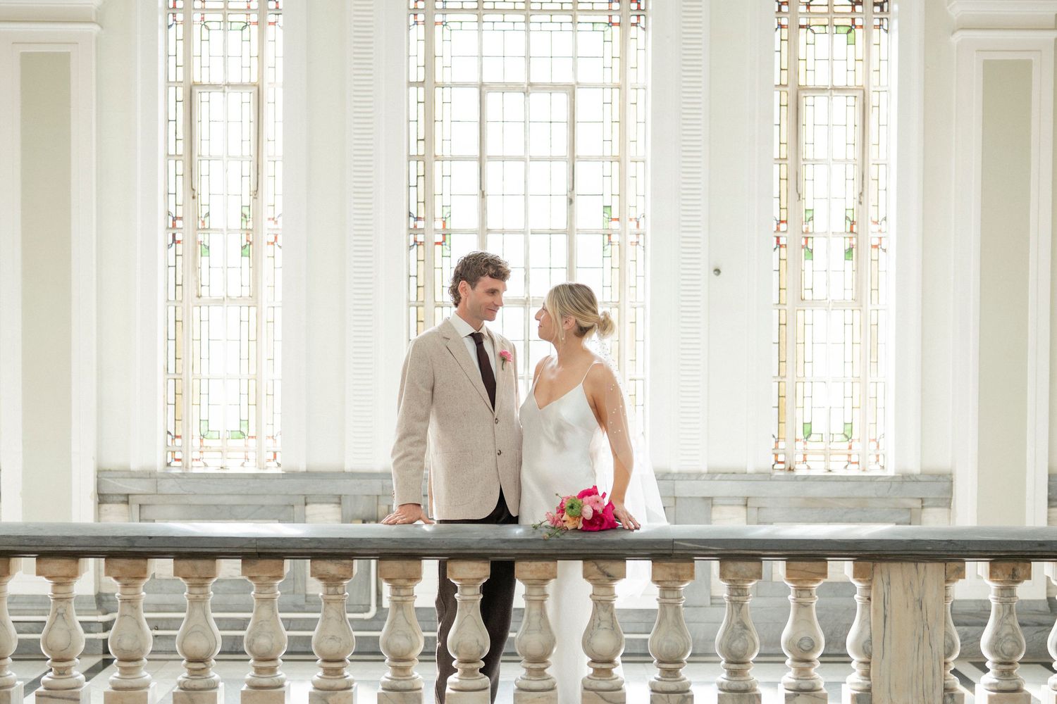 A bride in a white gown holding a pink bouquet stands beside a groom in a beige blazer on a balcony with tall arched windows.