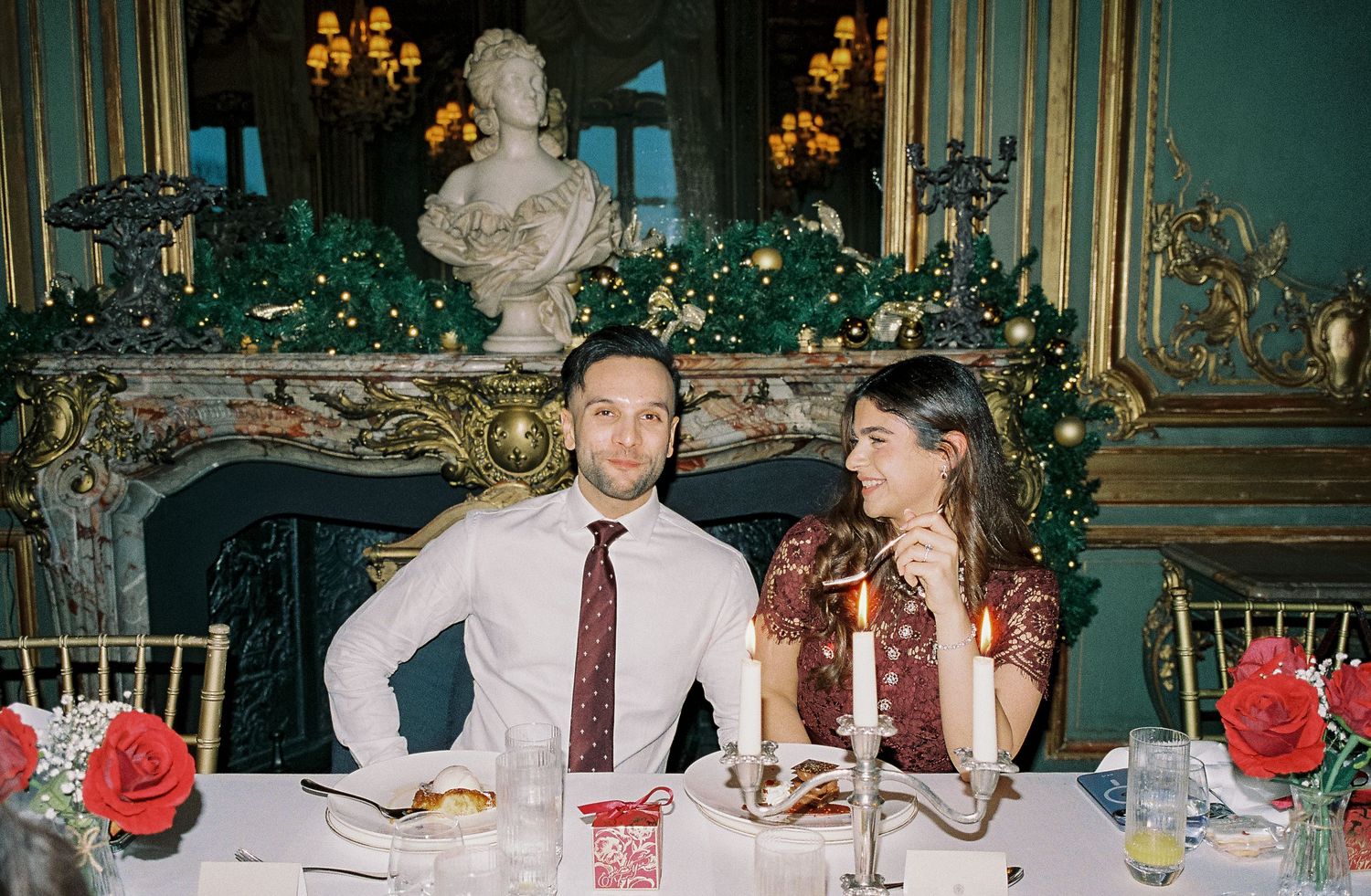 Couple sitting at elegant holiday dinner table with candles, red roses, and festive fireplace decor in ornate room.