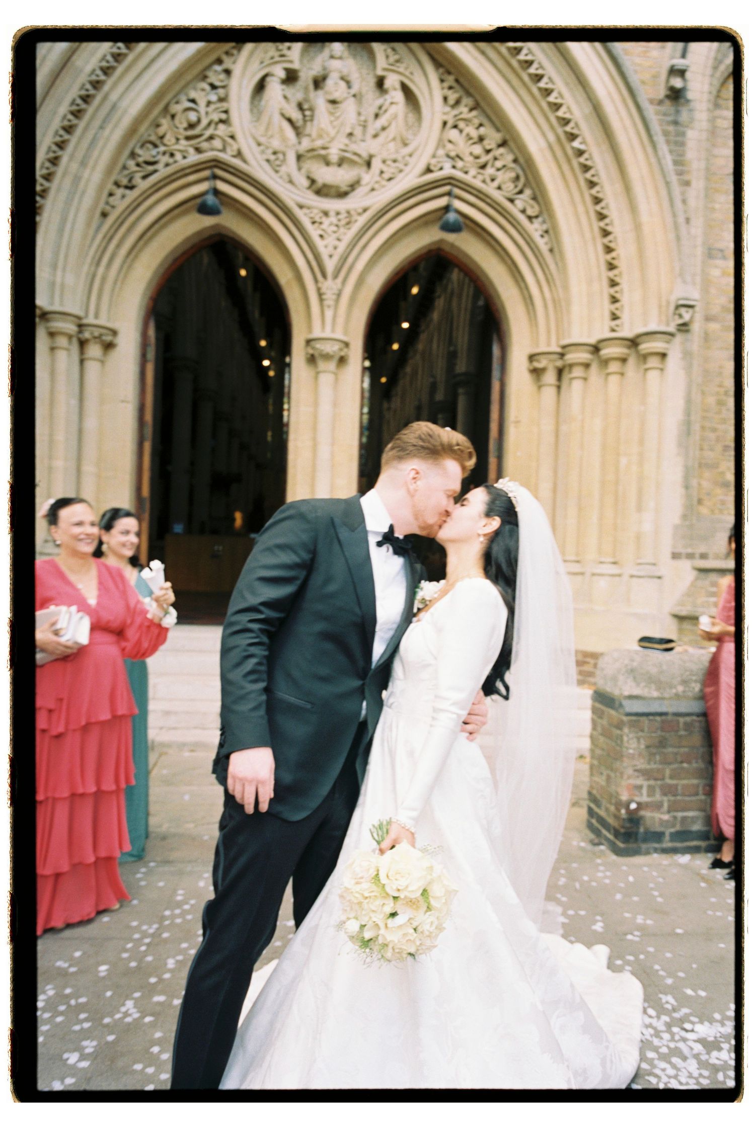 Bride and groom share a kiss outside gothic church entrance with wedding guests celebrating.