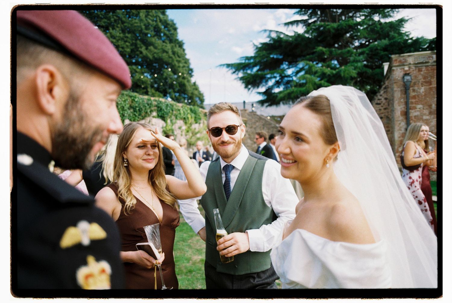 Wedding guests smiling and chatting outdoors, including a bride in white veil and a man in military uniform.