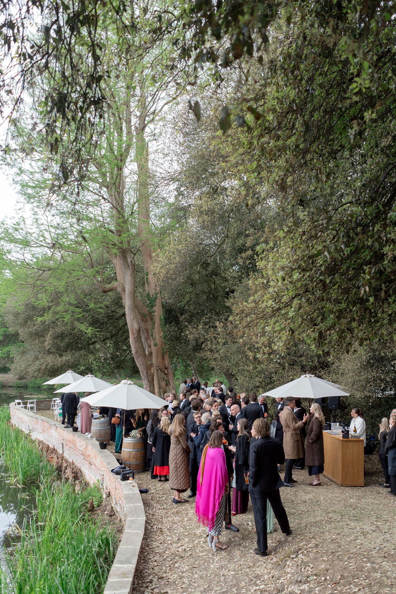 Wedding guests in elegant attire mingle beneath white umbrellas along a gravel path by a tranquil pond surrounded by tall trees.