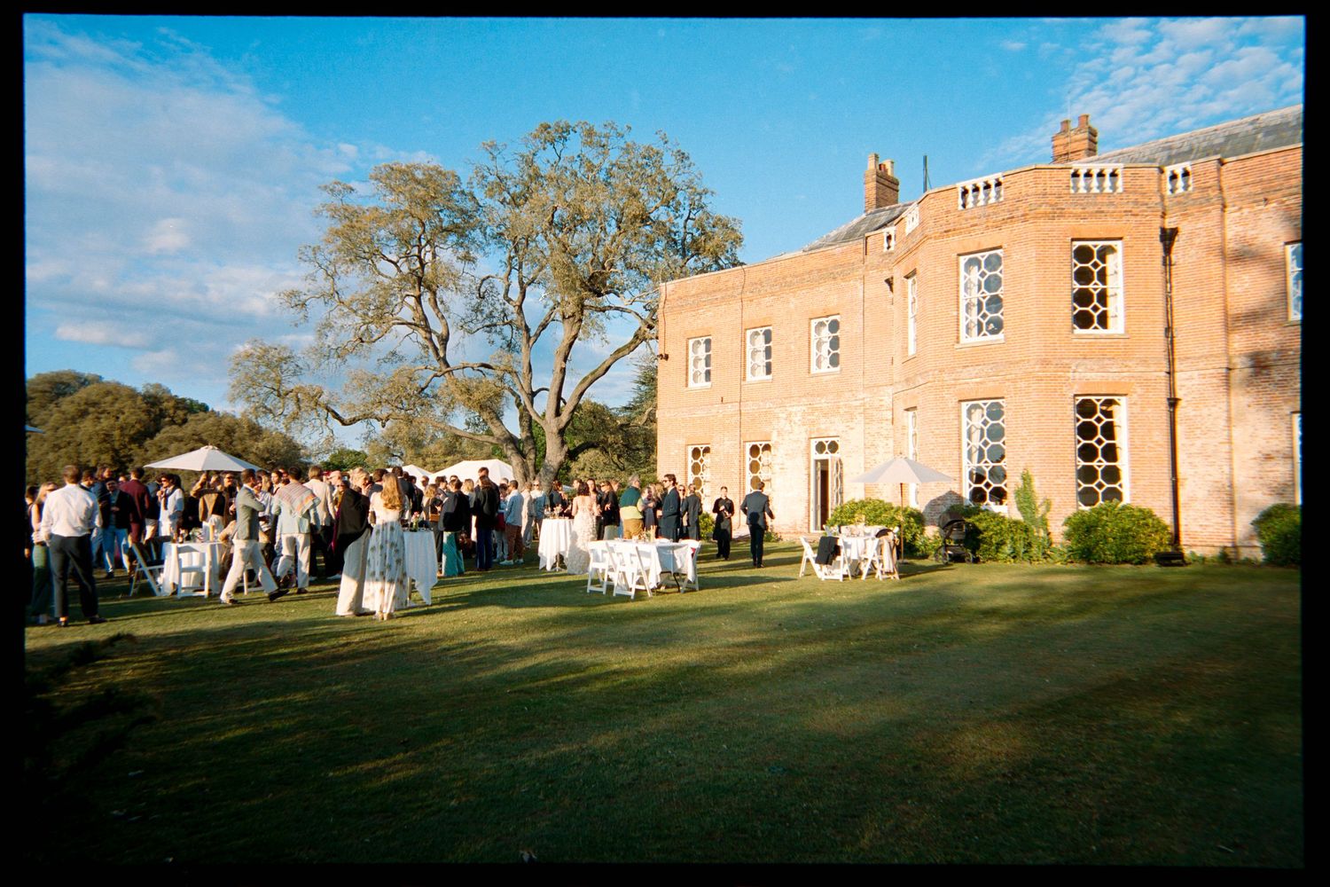 Wedding guests gather at white-draped tables on a sunlit lawn outside an elegant red brick manor house with tall windows and mature trees.