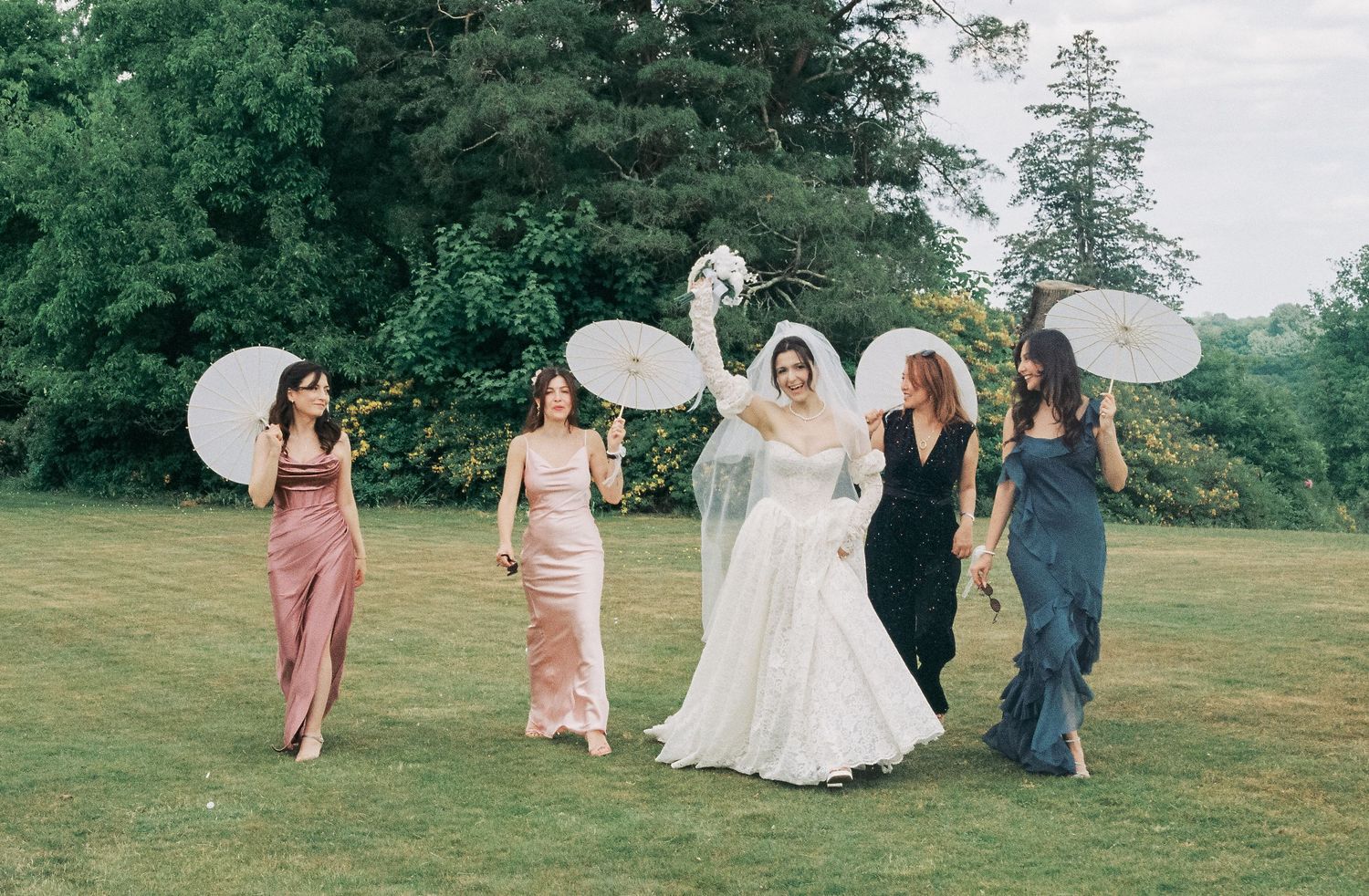 A bridal party in pastel and dark dresses poses with parasols on a lush green lawn during an outdoor celebration.