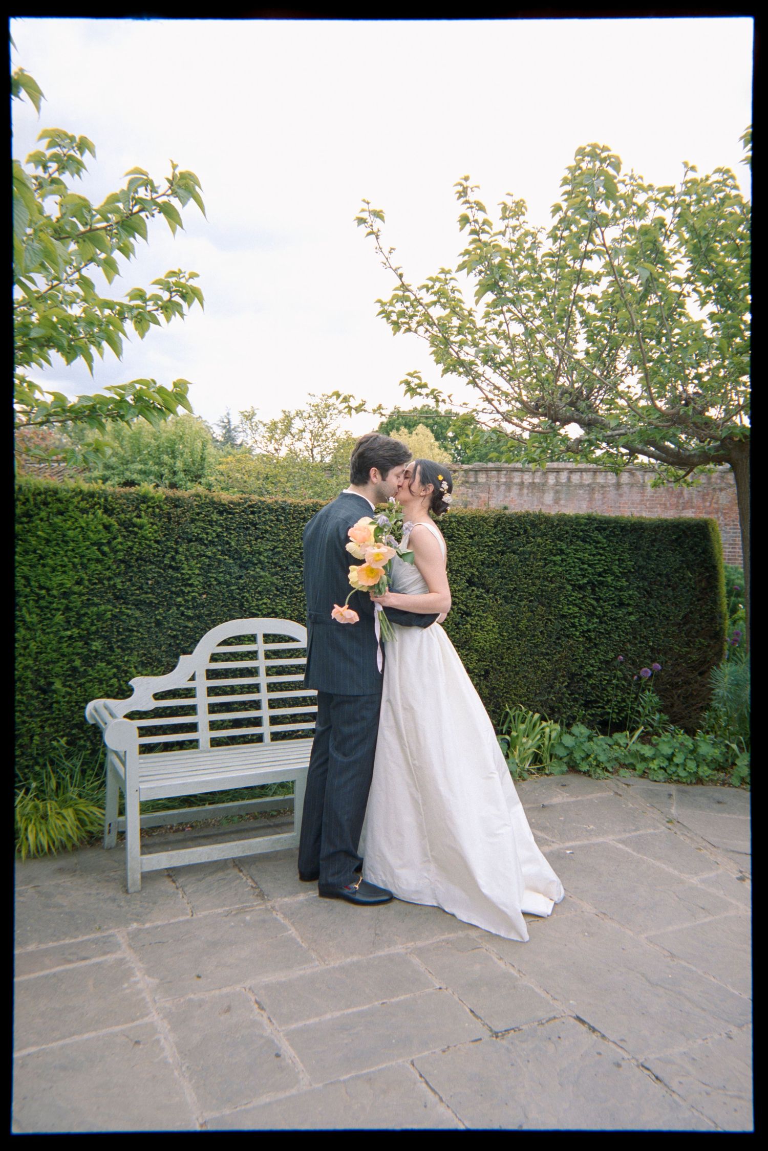 A bride in a white wedding gown and a groom in a dark suit kiss on a stone patio beside a bench and green hedge.