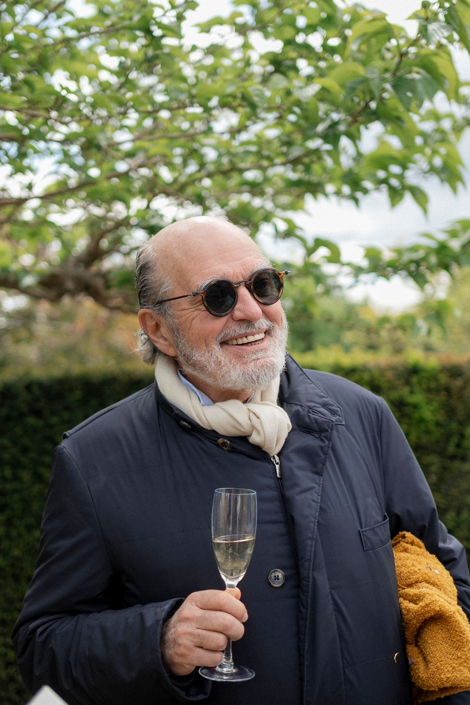 Smiling bearded man in round sunglasses and a navy jacket holds a glass of champagne in a leafy garden.