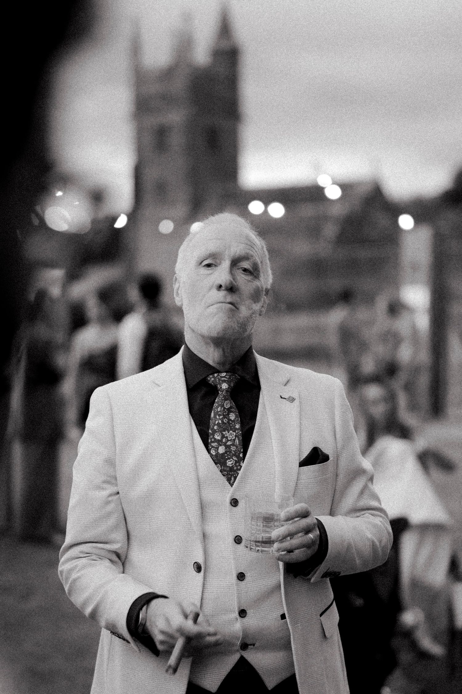 Distinguished older male wedding guest in white three-piece suit holding drink at evening outdoor reception.
