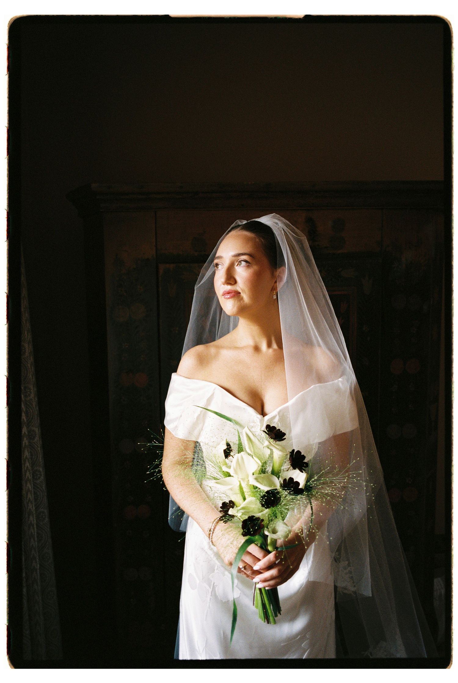 Bride in off-shoulder white gown holds bouquet of calla lilies and dark flowers, bathed in dramatic light.