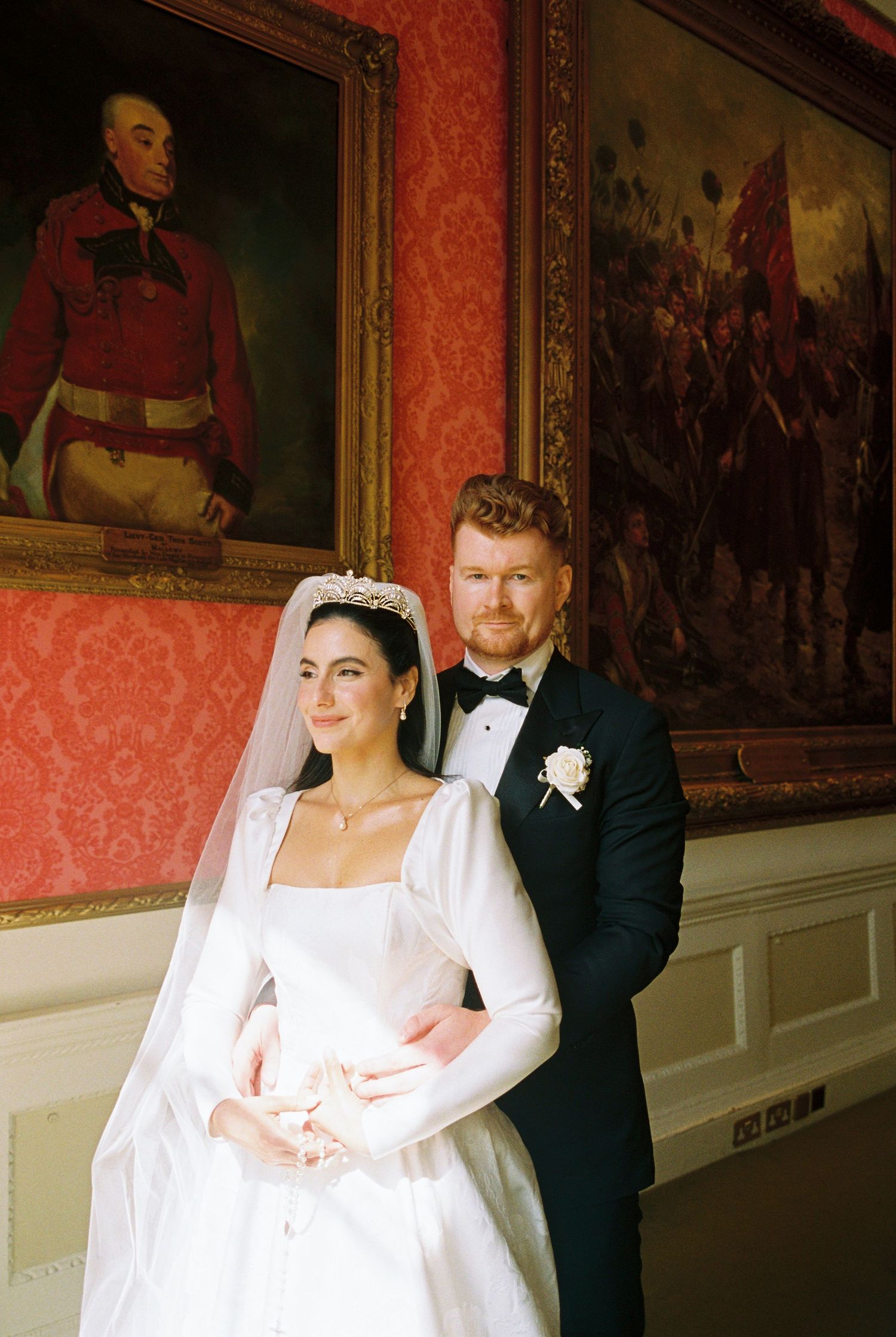 Elegant bride in white gown with tiara and groom in black tuxedo posing before ornate red wall paintings.
