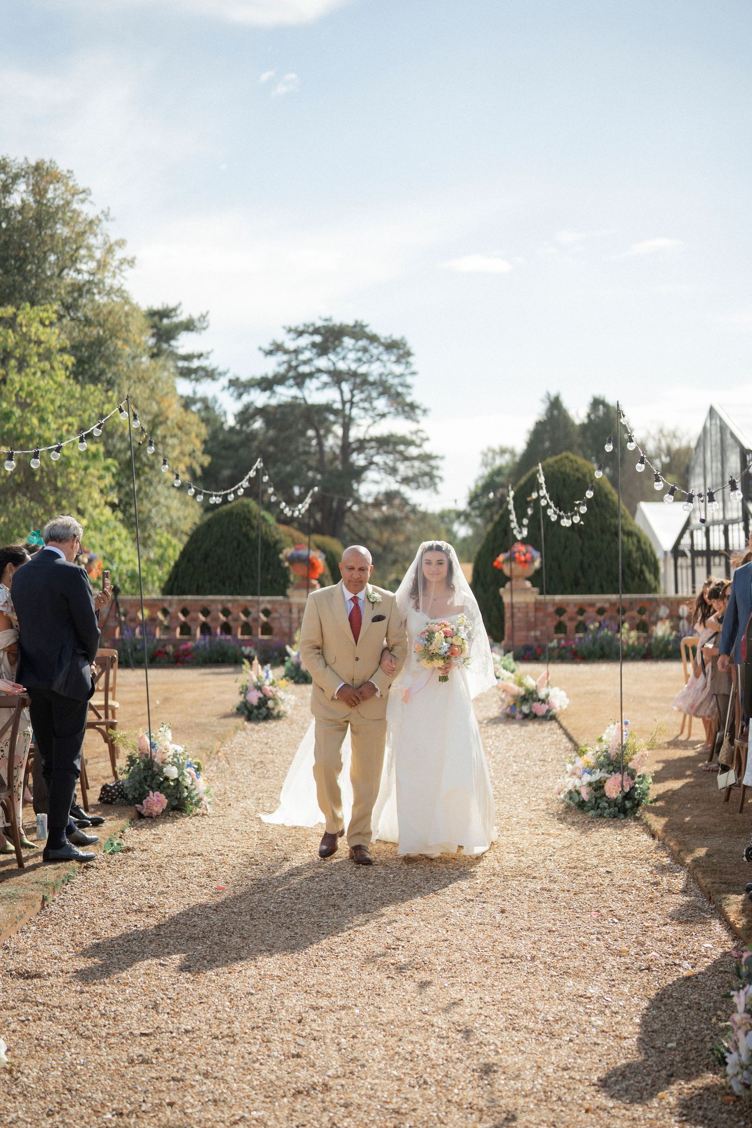 Bride walks down outdoor aisle with father at sunny garden wedding ceremony.