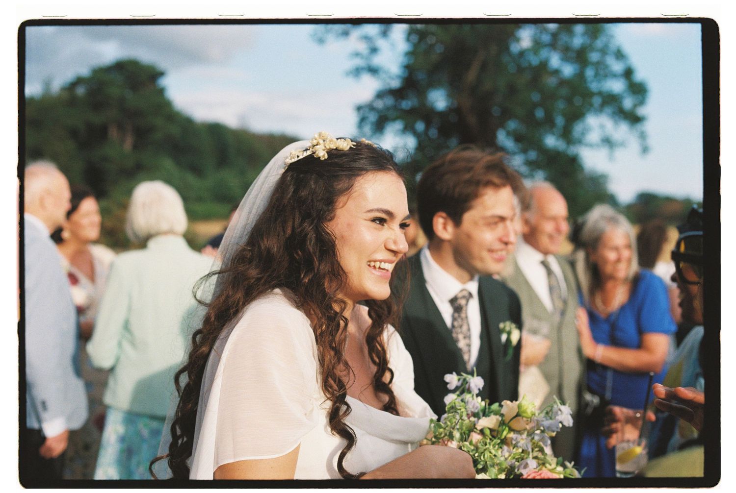 Smiling bride in white dress and floral headpiece holds bouquet beside groom at outdoor wedding ceremony.