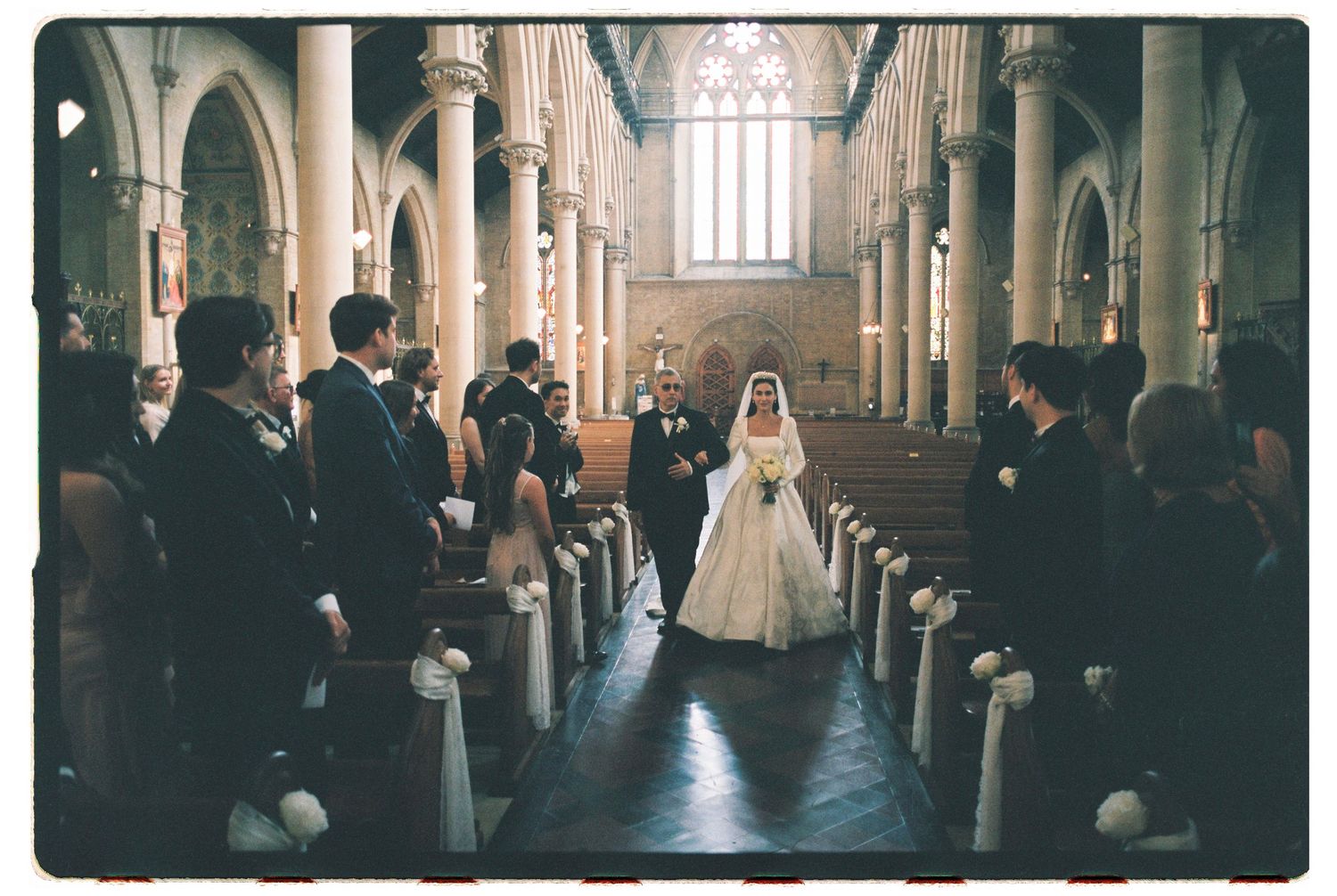 Bride in a white gown walks down the aisle of a grand church toward the groom as guests in dark attire watch from the pews.