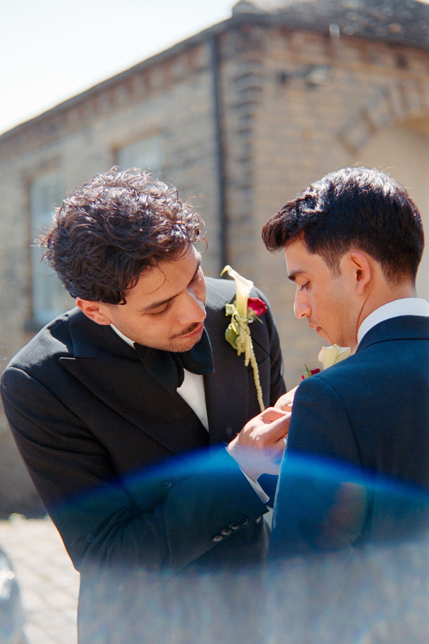 Two men in suits share an intimate moment outdoors against a stone building backdrop.