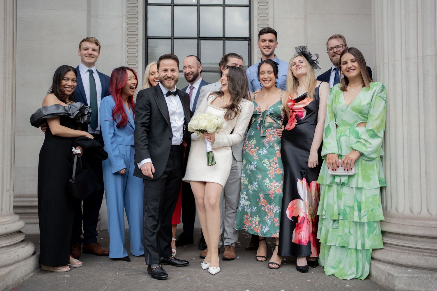 Wedding guests line up along a white columned building exterior for a group photo during a celebration.