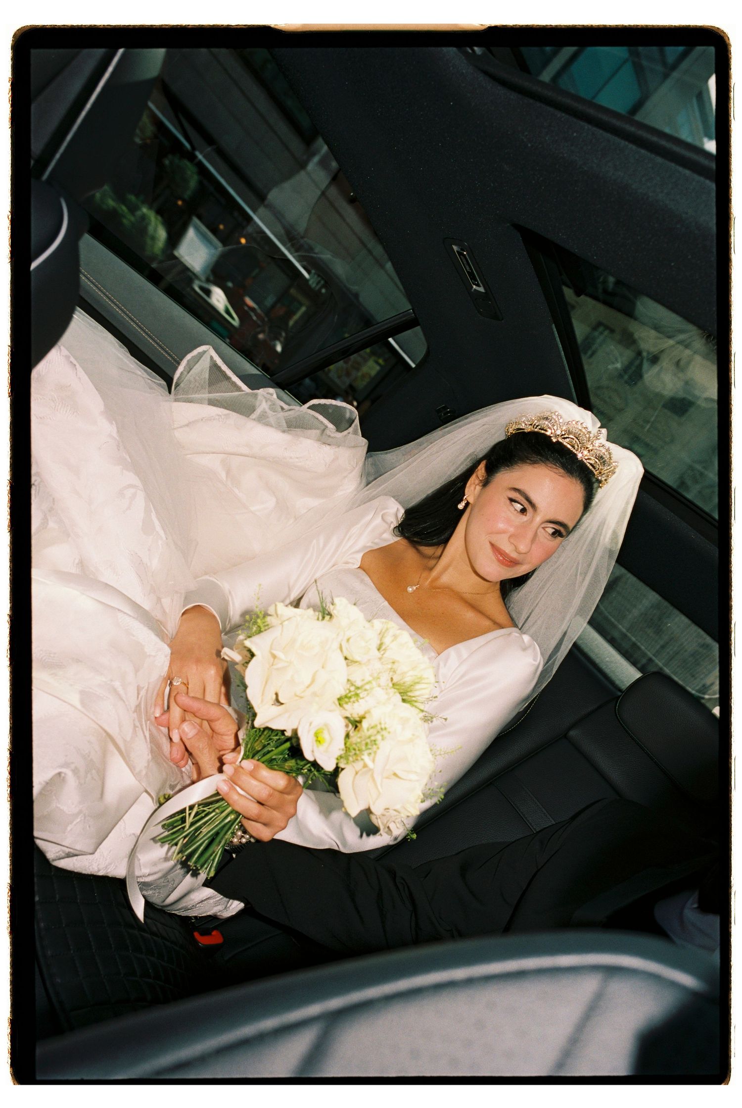 A bride in a white gown, tiara and veil sits in a car holding a bouquet of white roses and greenery.