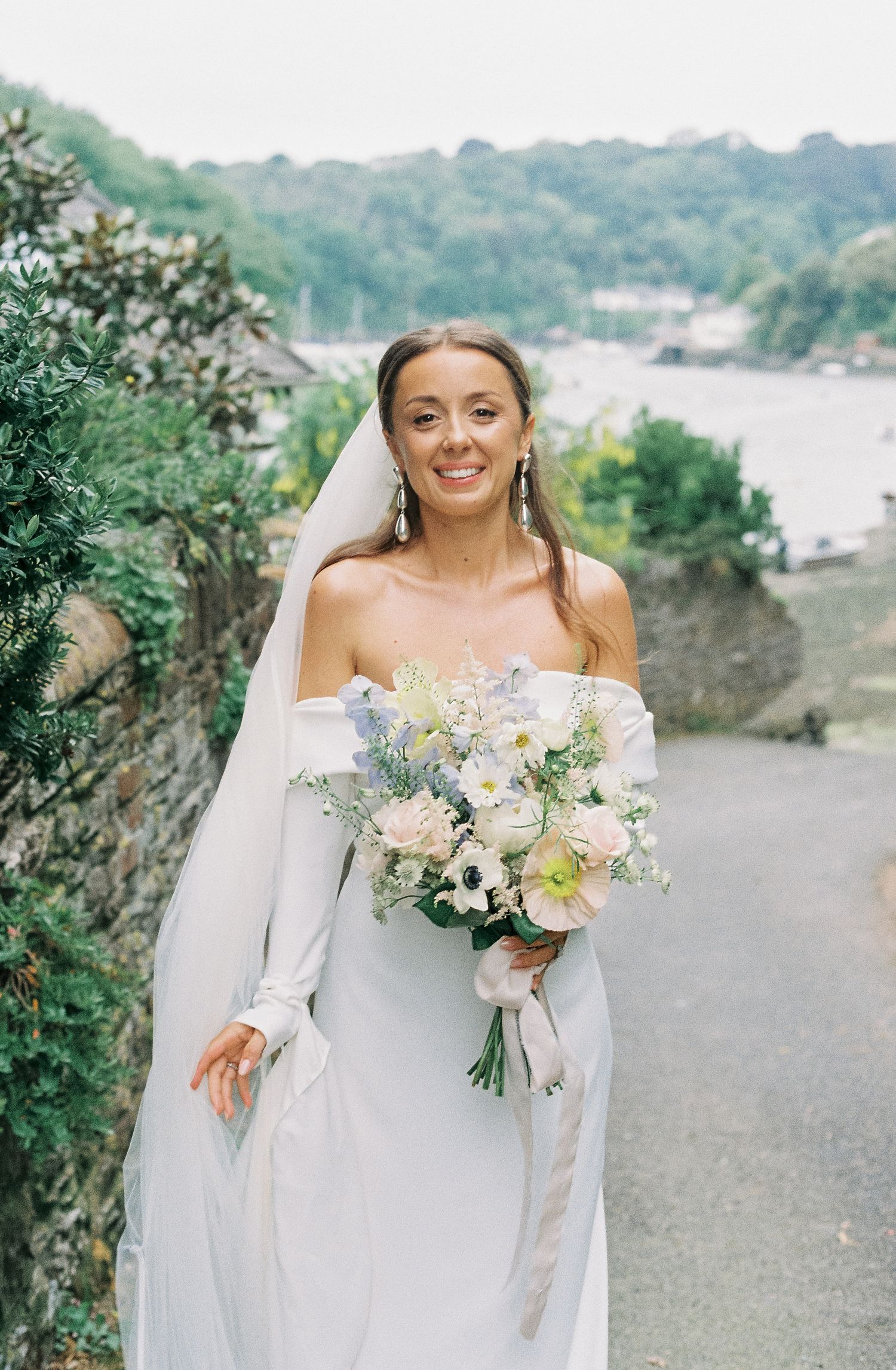 A bride in an off-shoulder white dress holds a delicate bouquet of flowers against a scenic coastal backdrop.