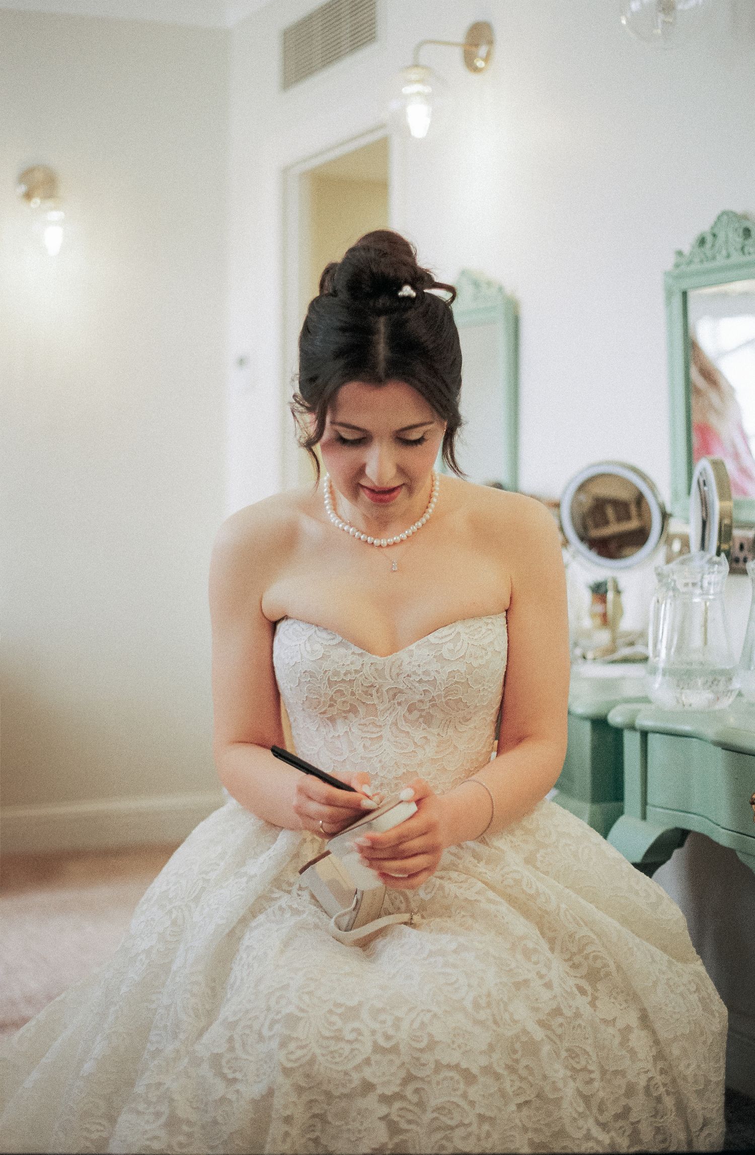 Bride in detailed lace gown sitting and reading note in bridal suite.