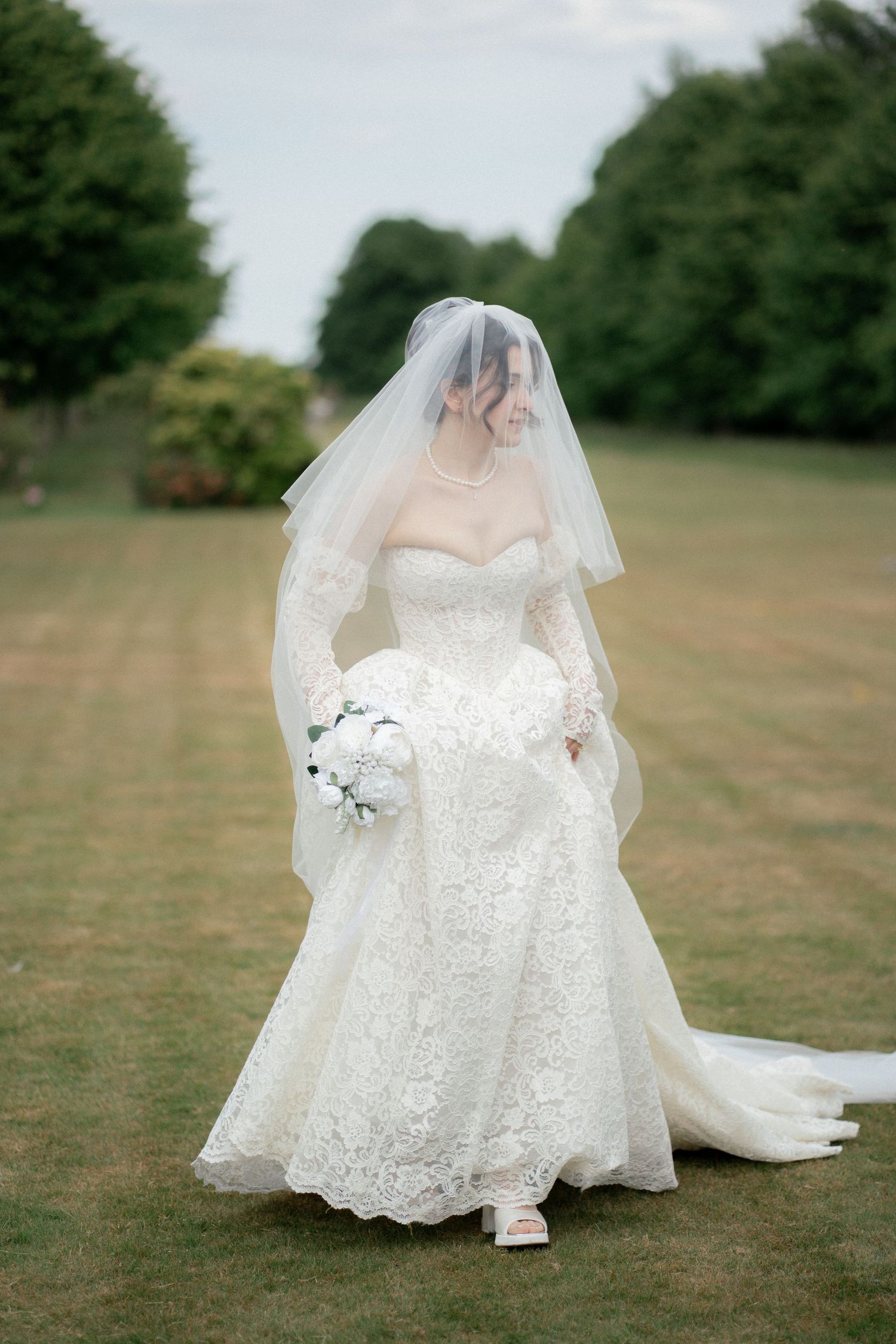 A flowing white lace wedding dress with delicate details and long veil spread out on green grass.