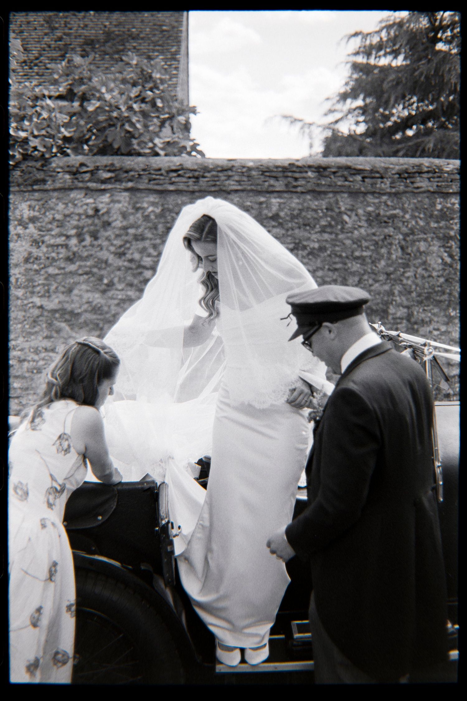 Black and white photo of a bride in white gown and veil being helped from a vintage car by a chauffeur.