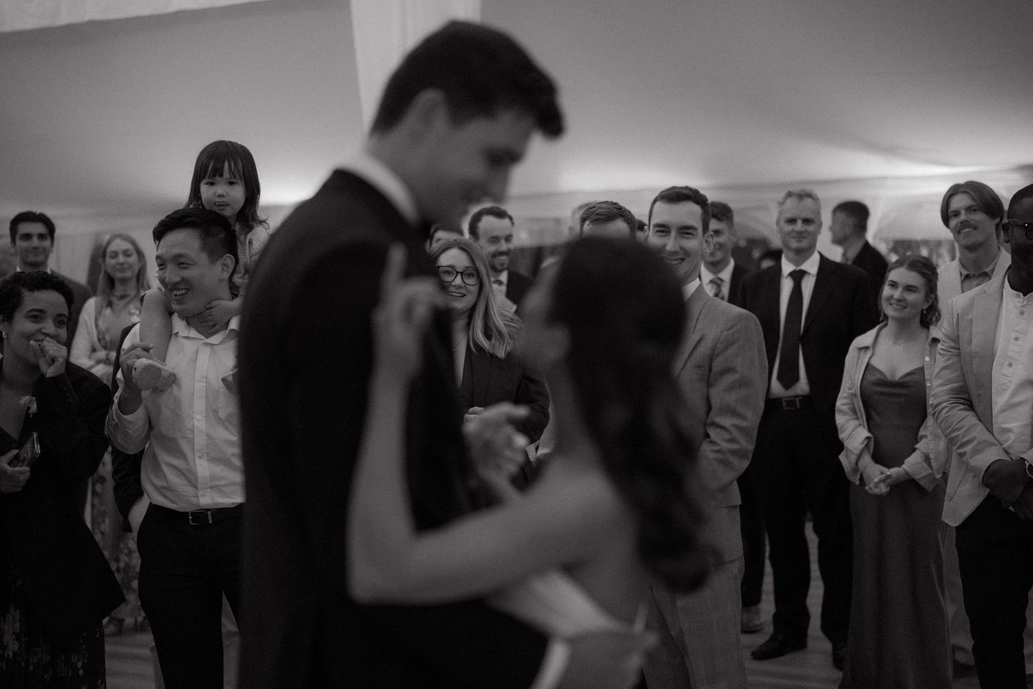 A couple shares their first dance at a wedding reception while guests gather around to watch in black and white.