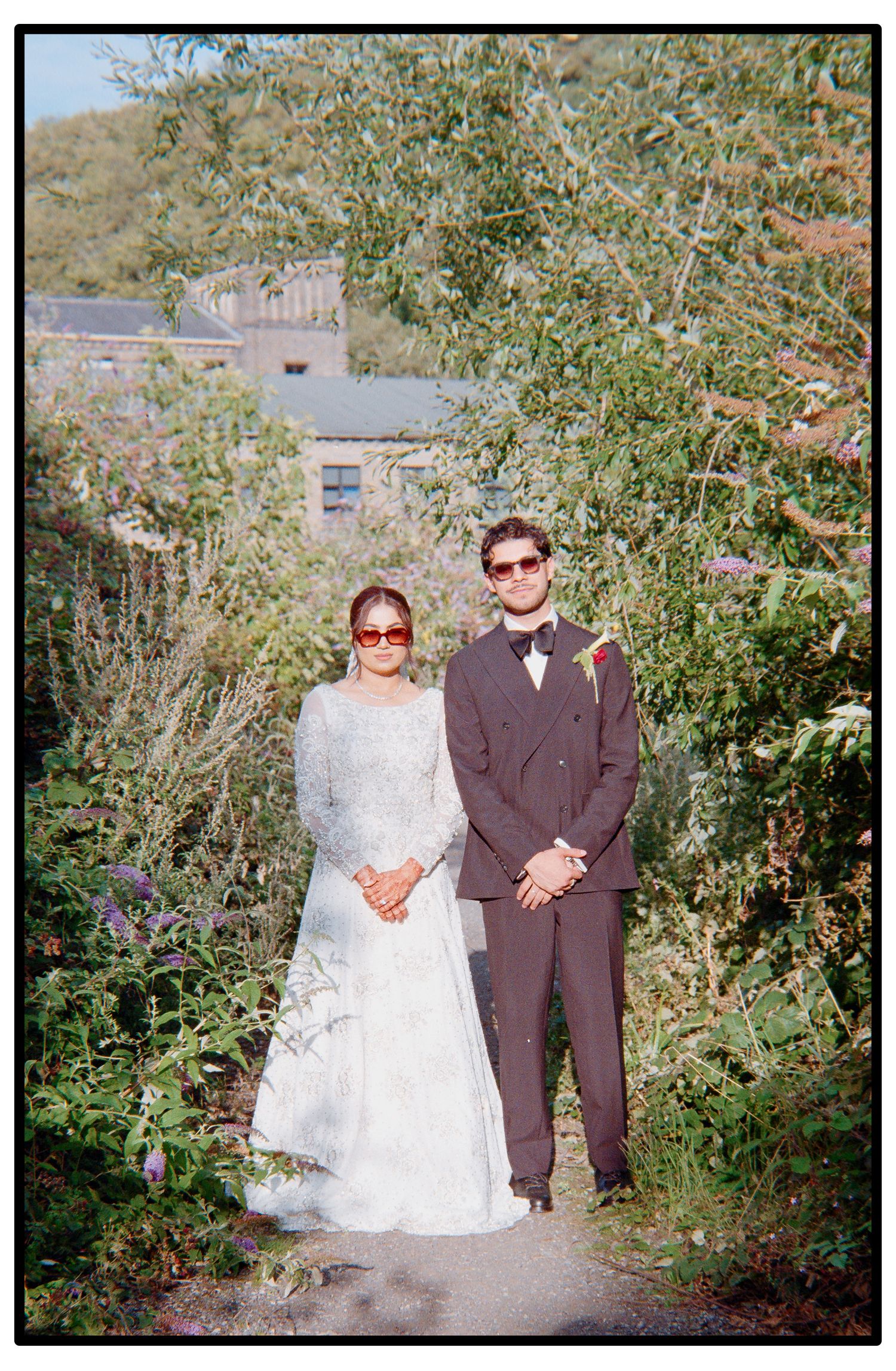 A couple poses together outdoors in formal wedding attire surrounded by green foliage on a sunny day.