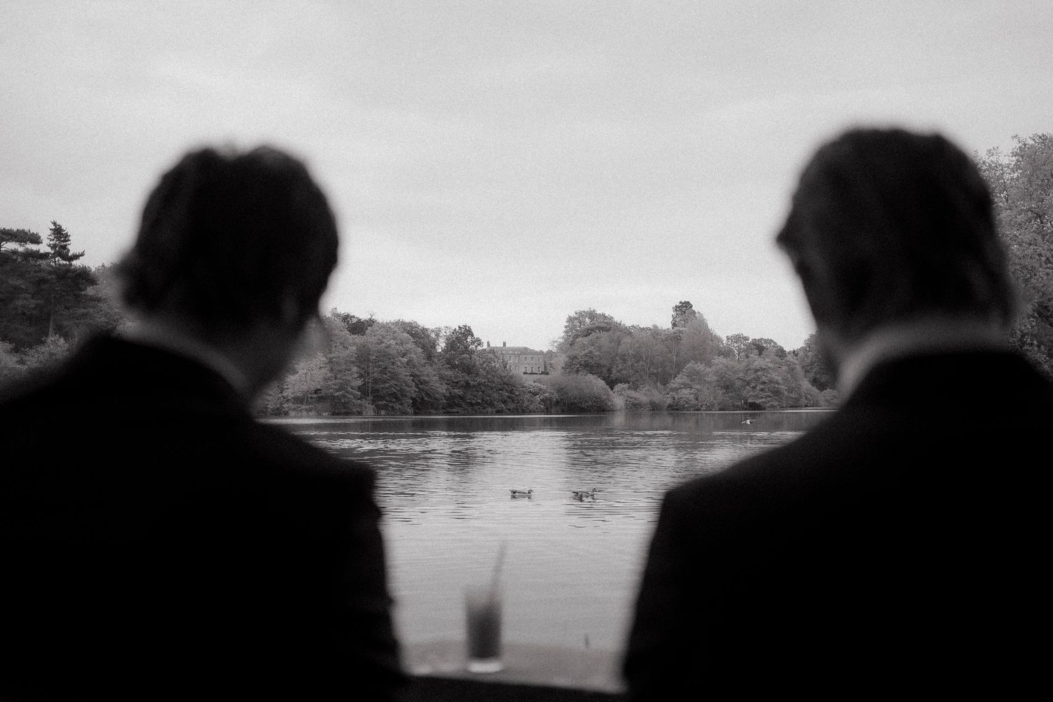 Black and white silhouettes of two men in suits look out over a quiet lake with ducks and trees, a drink on the ledge.